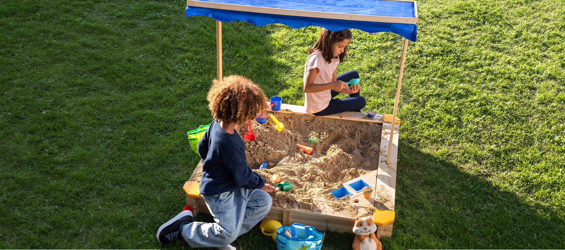 Zwei Kinder spielen in einem Holzsandelkasten mit blauem Sonnendach, umgeben von grünem Gras.