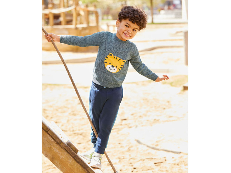 Ein Junge trägt einen blauen Pullover mit Tigermotiv und blaue Hose auf einem Spielplatz.