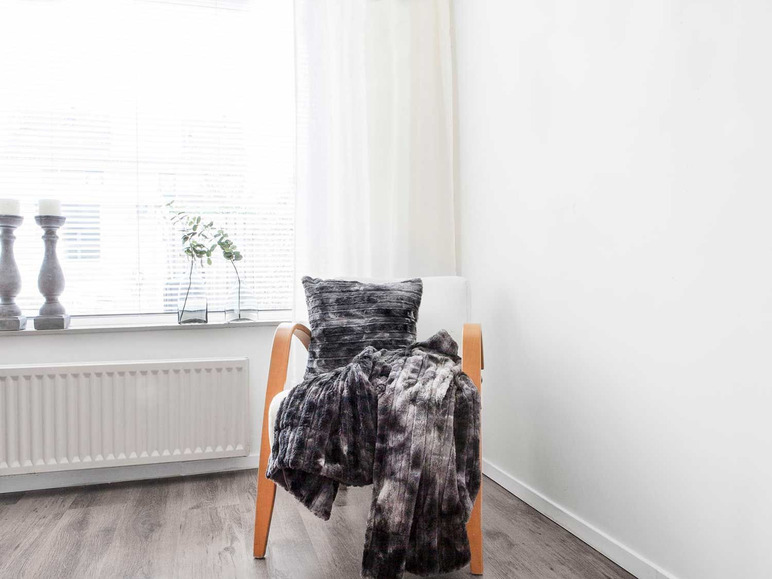 Ein Sessel mit einer grauen Decke und einem Kissen, vor einem Fenster platziert.