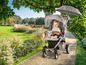 Ein Baby in einem grauen Kinderwagen mit Sonnenschirm auf einem Weg in einem Garten.