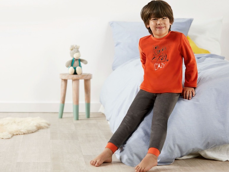 Junge in orangem Pyjama mit grauer Hose auf einem Bett.