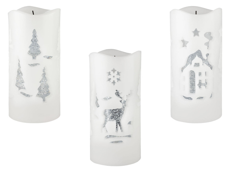 Drei weiße Weihnachtskerzen mit silbernen Glitzermotiven von Tannenbäumen, einem Hirsch und einem Haus.
