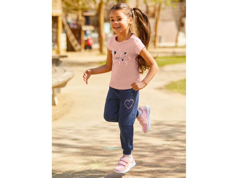 Ein Mädchen trägt ein rosa T-Shirt mit Herzchen-Druck und blaue Jogginghose.