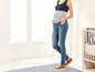 Schwangere Frau in Jeans und Tanktop, stehend vor einem Babybett.