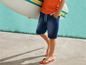 Ein Junge in Jeanshorts und Flip-Flops mit einem Surfbrett.