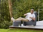 Ein Mann sitzt auf einem grauen Loungesessel und einem passenden Hocker auf einer Holzterrasse.