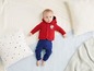 Baby in roter Jacke und blauer Hose liegt auf einem weißen Bett.