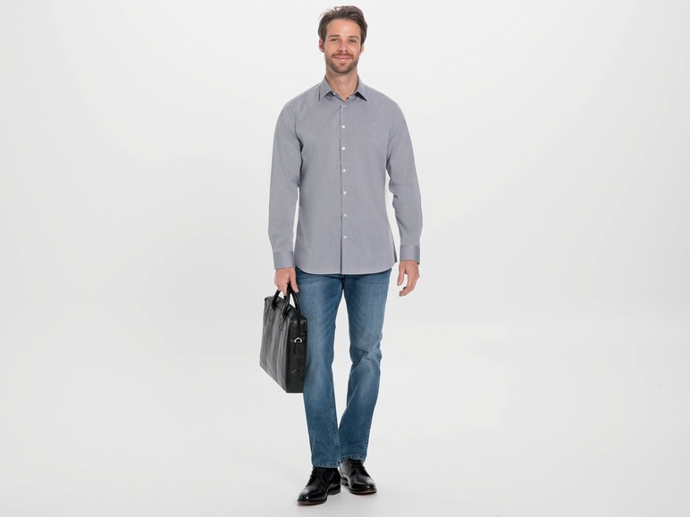 Mann in einem grauen Hemd und blauen Jeans mit einer schwarzen Laptoptasche.