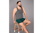 Mann in einem grauen Tanktop und grünen Boxershorts sitzt auf einem Holzhocker.