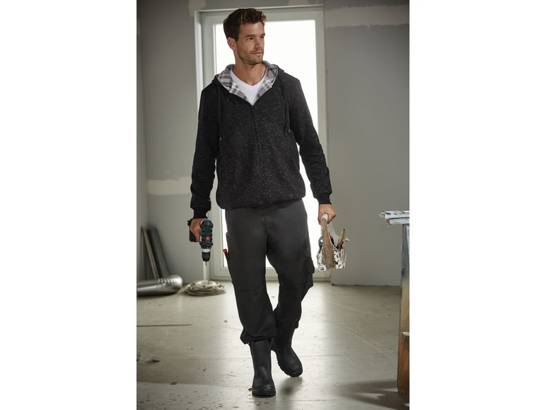 Mann in schwarzem Hoodie und Arbeitskleidung mit einer Bohrmaschine und einer Werkzeugkiste.