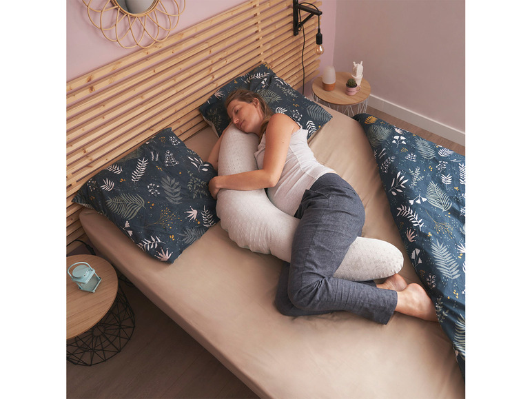 Eine Frau schläft auf der Seite mit einem Schwangerschaftskissen und einem Kissen mit botanischem Muster.