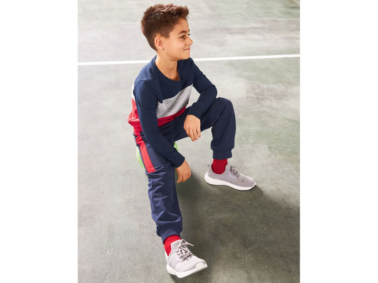 Junge in blauen Jogginghose und Pullover mit roten Streifen, trägt graue Sneaker.
