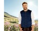 Mann mit blauer Weste und Lederhosen auf einer Wiese.