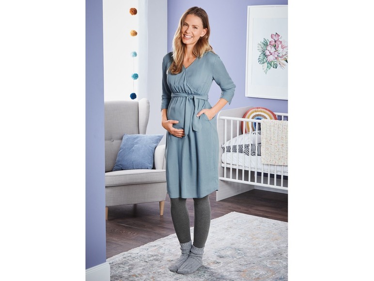 Eine schwangere Frau in einem blauen Kleid und Strumpfhosen in einem Babyzimmer.