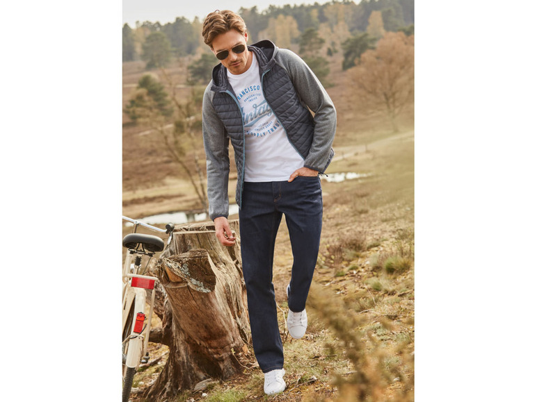 Mann in einer grauen Jacke, weißem T-Shirt und blauen Jeans.