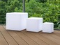Drei weiße, kubische LED-Hocker auf einer Holzterrasse.