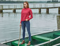 Eine Frau in einem roten Pullover und blauen Jeans steht auf einem Boot am See.
