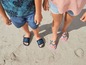 Zwei Kinder in Sandalen am Strand, eines mit NYC-Druck und das andere mit Regenbogenmuster.