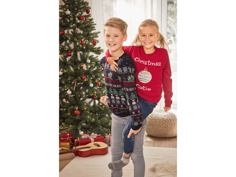 Zwei Kinder in Weihnachtspullovern vor einem Weihnachtsbaum.