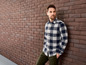 Mann in einem blau-weiß karierten Hemd steht vor einer Backsteinwand.