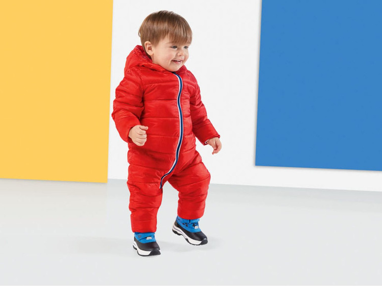 Ein kleiner Junge in einem roten Winteroverall und blauen Schuhen.