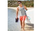 Mann in grauer Tanktop, roten Badehosen und Flip-Flops am Strand mit einem Paddleboard.