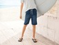 Ein Junge trägt Jeans-Shorts und Flip-Flops am Strand mit einem Surfbrett.