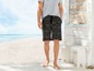 Mann in schwarzen Shorts mit tropischem Muster am Strand.