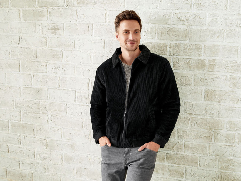 Ein Mann trägt eine schwarze Cordjacke und graue Jeans vor einer weißen Backsteinwand.