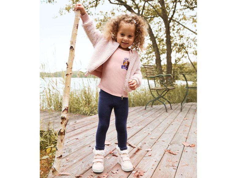 Ein Mädchen trägt einen rosa Pullover mit Paw Patrol-Aufdruck, blaue Leggings und weiße Sneaker.