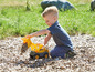 Ein Junge spielt mit einem gelben Spielzeugbagger auf einem Spielplatz mit Holzspänen.