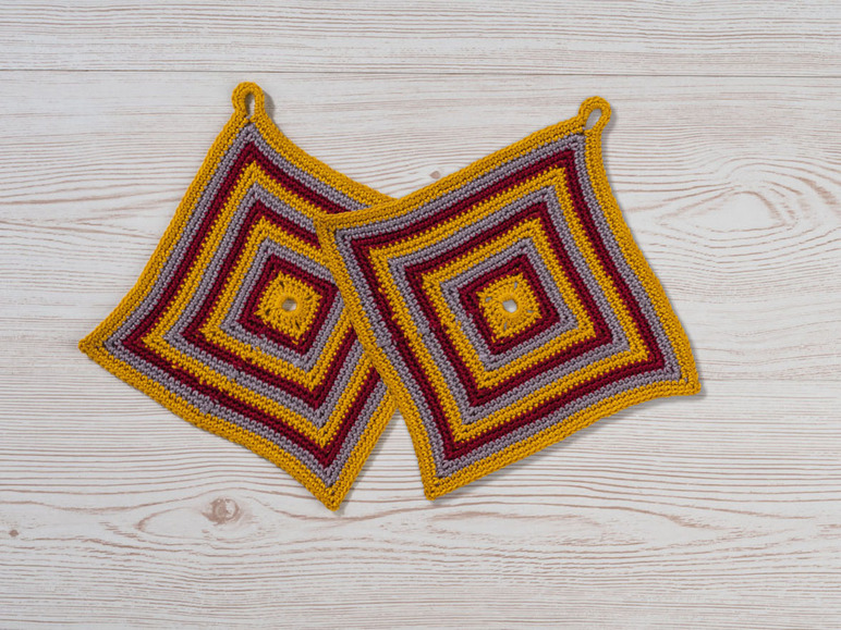 Zwei gehäkelte Topfhandschuhe in Gelb, Grau und Rot auf einem Holztisch.