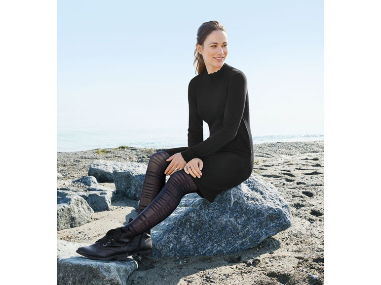 Eine Frau in einem schwarzen Kleid und Strumpfhosen sitzt auf einem Felsen am Strand.