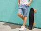Ein Junge in Jeans-Shorts und weißen Sneakers lehnt an einer türkisfarbenen Wand mit einem Skateboard.