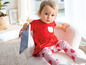 Ein Baby-Mädchen in einem roten Kleid mit Apfel-Print und rosa Hose mit Apfel-Print hält ein Buch.