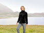 Eine Frau in einer schwarzen Fleecejacke und grauen Hosen geht in einer Landschaft mit Bergen.