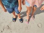 Zwei Kinder am Strand tragen Sandalen, ein Paar mit NYC-Logo und ein Paar mit Regenbogenmuster.