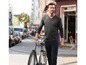 Mann in einem grauen Pullover und blauen Jeans geht mit einem Fahrrad auf der Straße.
