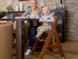 Ein Baby sitzt auf einem Hauck Holzhochstuhl am Tisch mit einem Erwachsenen.