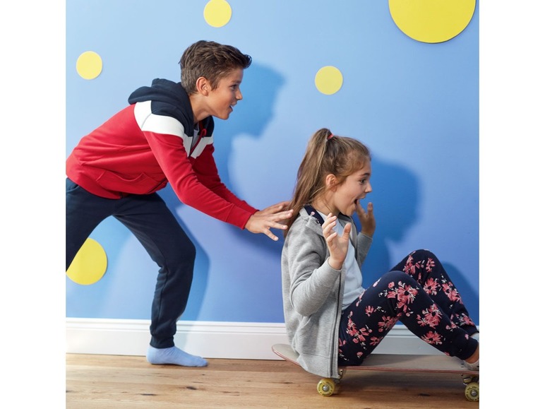 Ein Junge in einem roten Pullover schiebt ein Mädchen auf einem Skateboard.