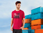 Ein Mann trägt ein rotes T-Shirt mit industriellem Print vor einem Stapel Container.