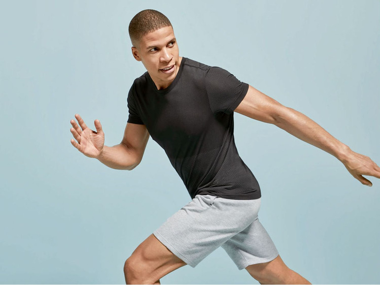 Mann in schwarzem T-Shirt und grauen Shorts posiert vor blauem Hintergrund.