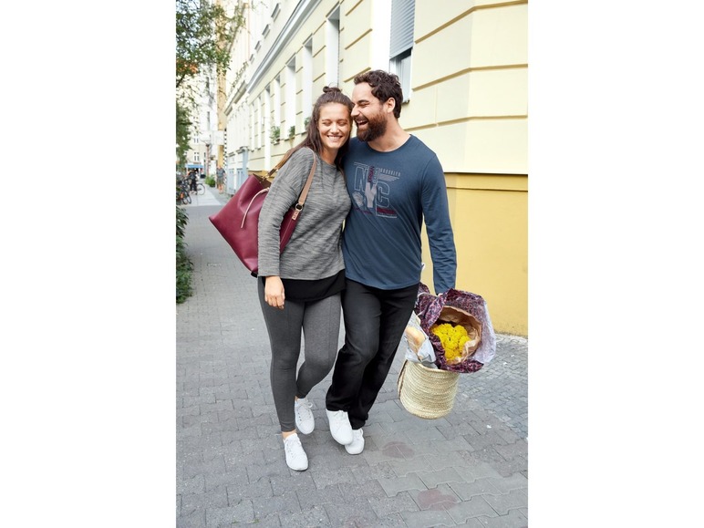 Ein Paar geht auf der Straße spazieren, die Frau trägt eine rote Tasche und der Mann trägt Blumen.