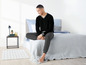 Ein Mann in schwarzer Pyjamahose und -pullover sitzt auf einem Bett.