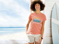 Frau in einem rosafarbenen T-Shirt mit einem Surfbus-Motiv und weißen Shorts, vor Strandkulisse.