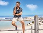 Mann in einem blau-weiß karierten Hemd und beige Shorts sitzt auf einem Geländer am Meer.