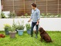 Ein Mann gießt Pflanzen mit einem Schlauch im Garten, neben ihm ein Hund.