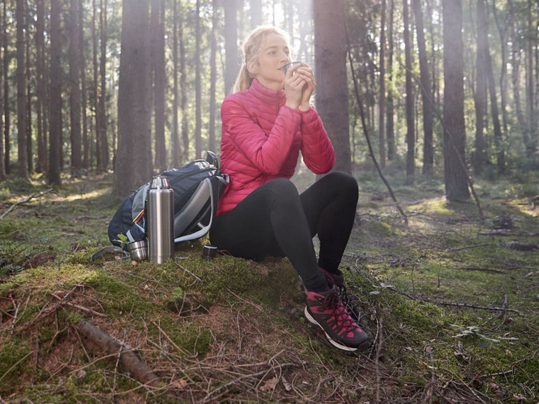 Eine Frau im Wald mit einer Thermoskanne und einem Rucksack.