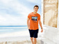 Mann in einem orangen T-Shirt mit 'Adventures Traveling'-Aufdruck und blauen Shorts.