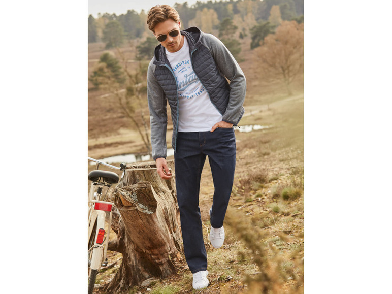 Mann trägt eine graue Jacke, ein weißes T-Shirt und blaue Jeans.